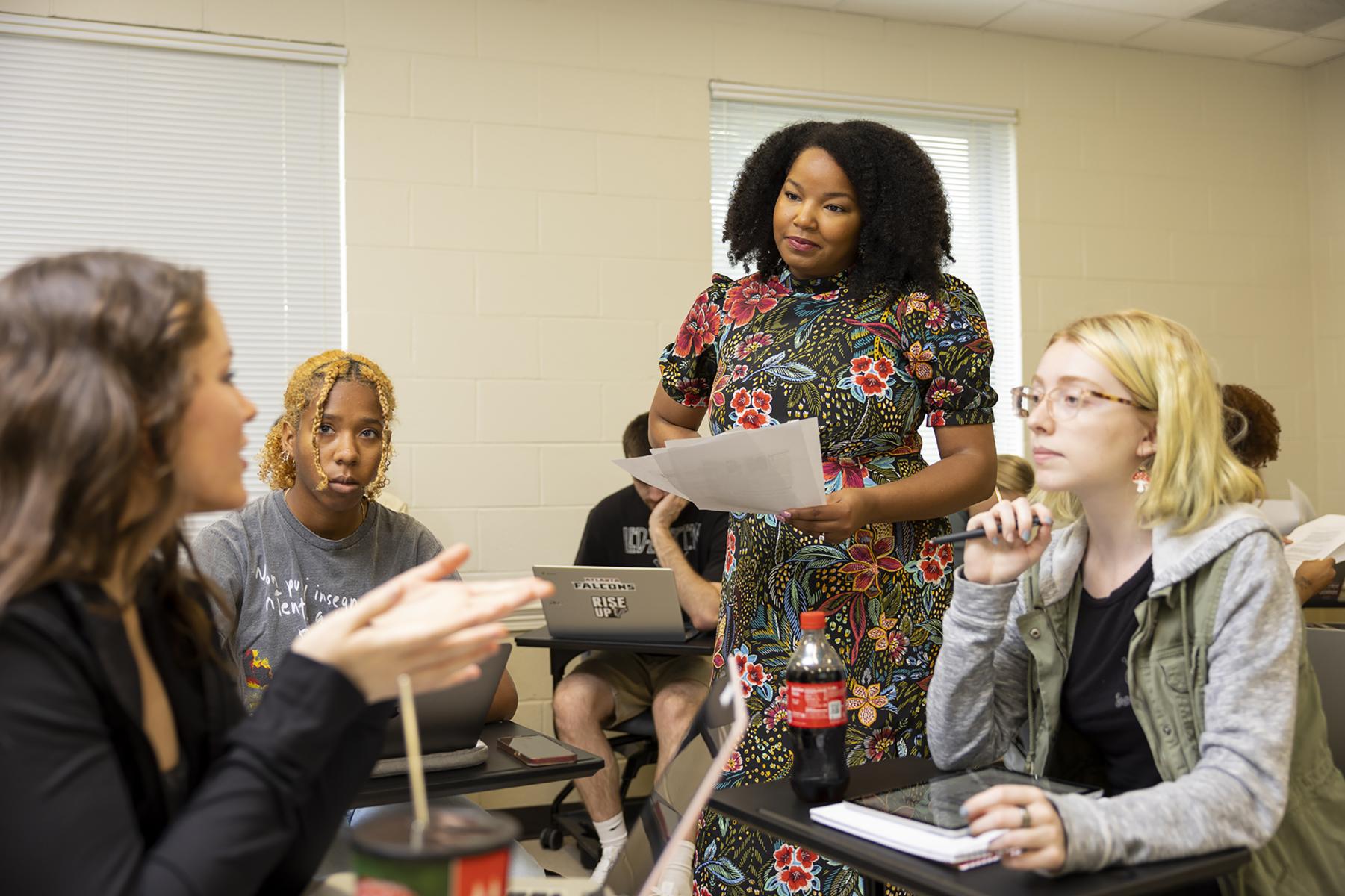 Dr. Johnson standing in her class amongst her students who are all sitting at their desks in a circle while one student has an active discussion with her. 